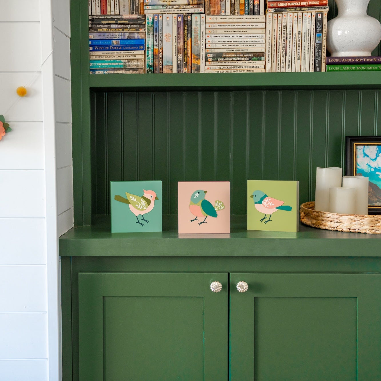 Green cabinet with decorative paper bird frames, styled near books, candles, and framed pictures in a room.