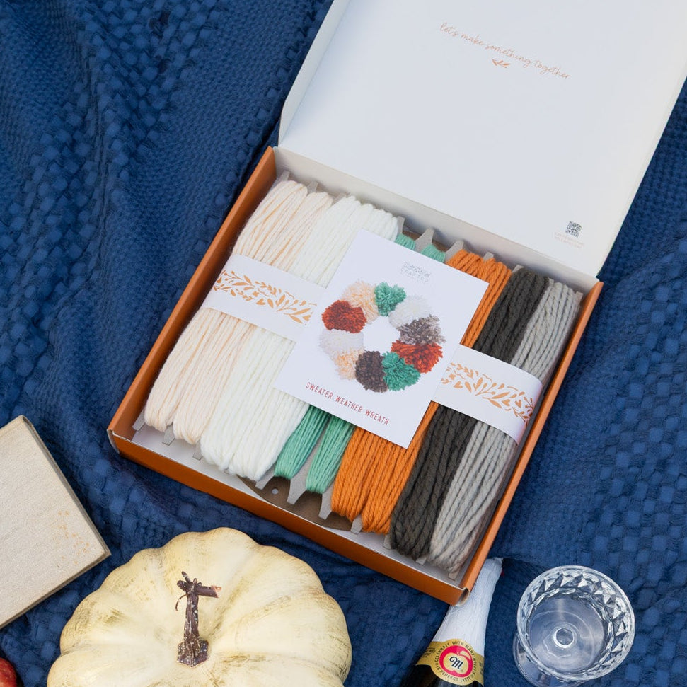 An outdoor picnic setup on a textured navy blue blanket, featuring a craft project kit filled with yarn, two clear drinking glasses, a bottle of cider, a small white pumpkin.