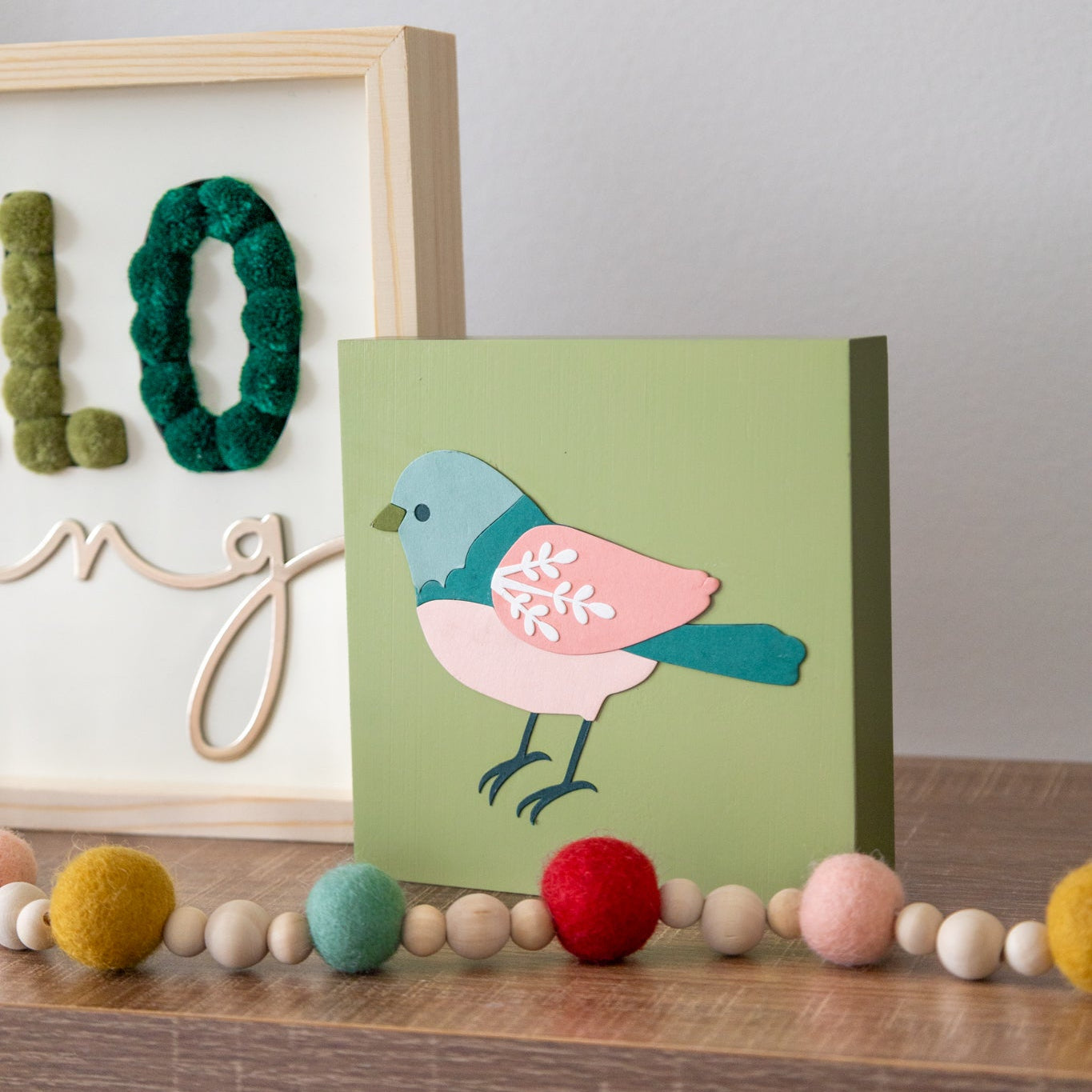 Decorative craft sign with a bird illustration on a shelf with colorful pom-poms.