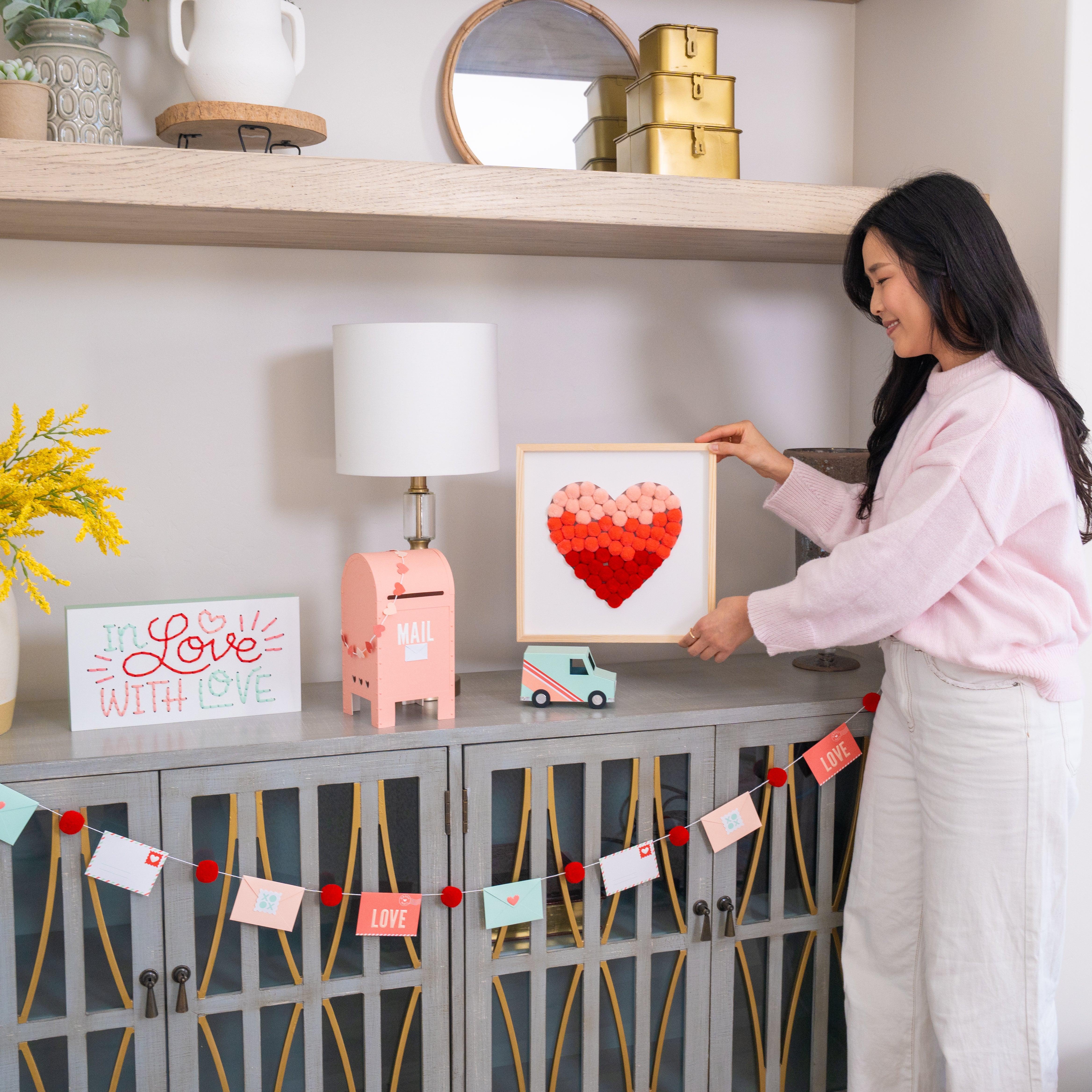 Woman arranging decor items on a shelf with a heart-shaped pom pom frame