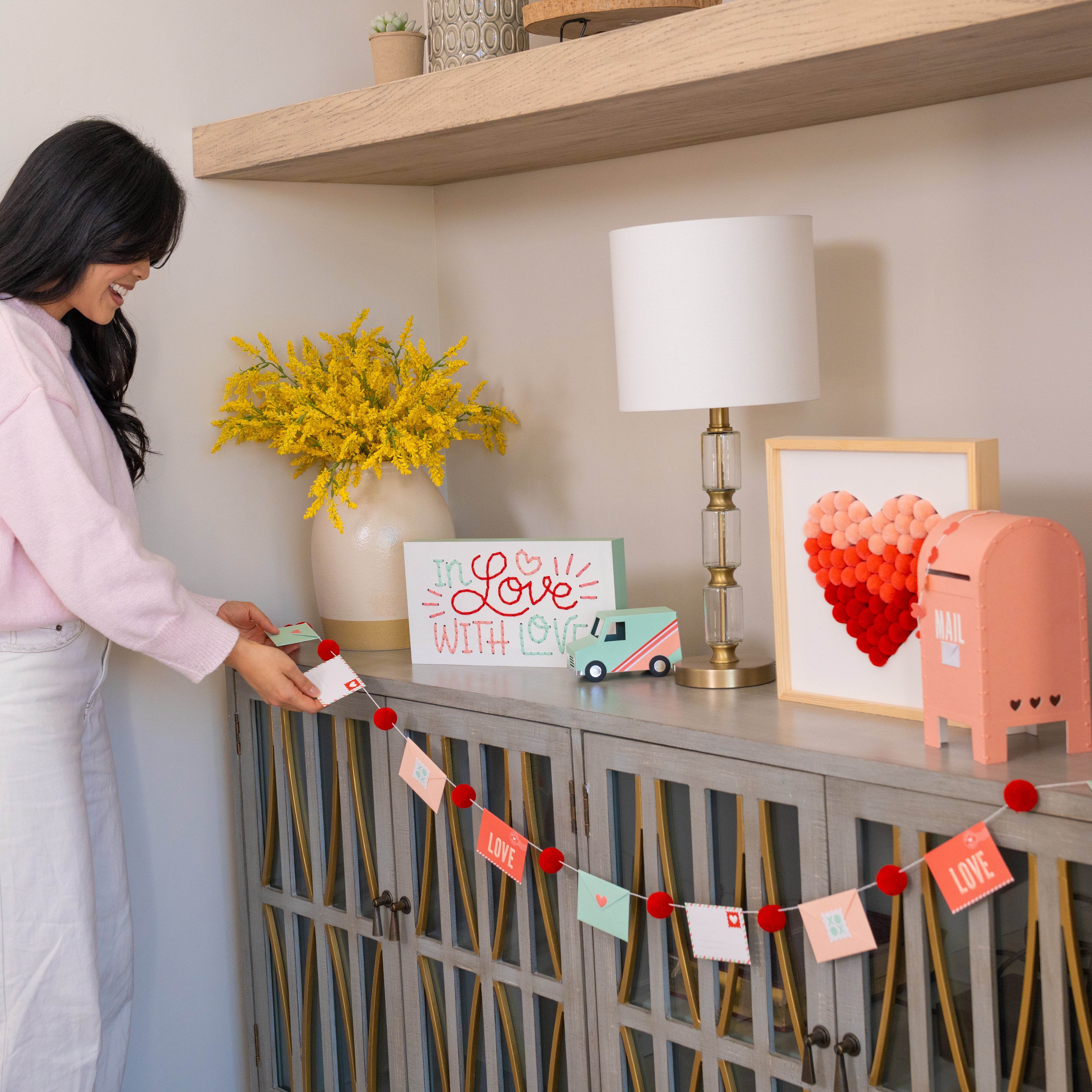 Woman standing next to a decorative cabinet with various crafted items including a love letter garland
