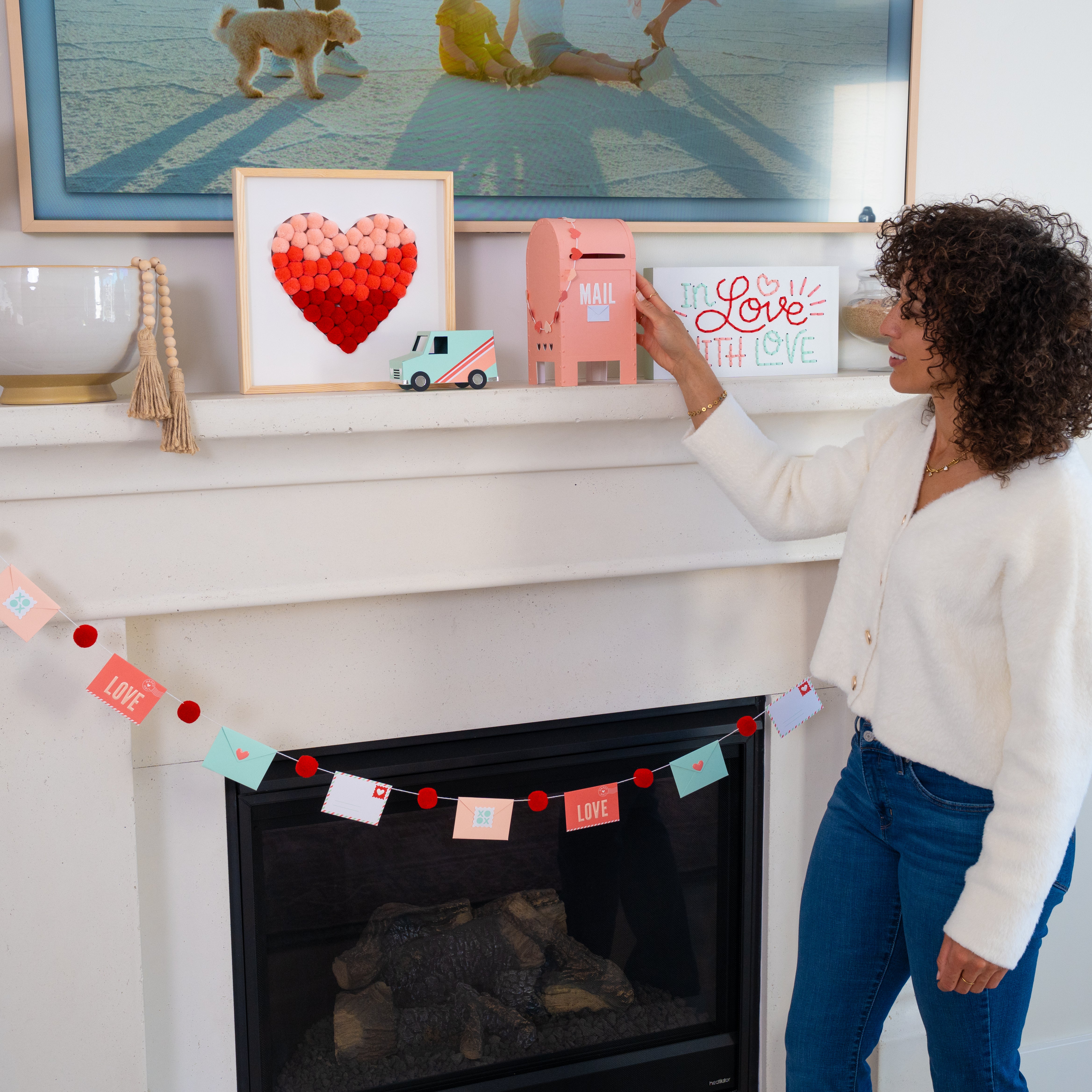 Woman placing a small pink mailbox on a fireplace mantle decorated with Valentine's decor items.