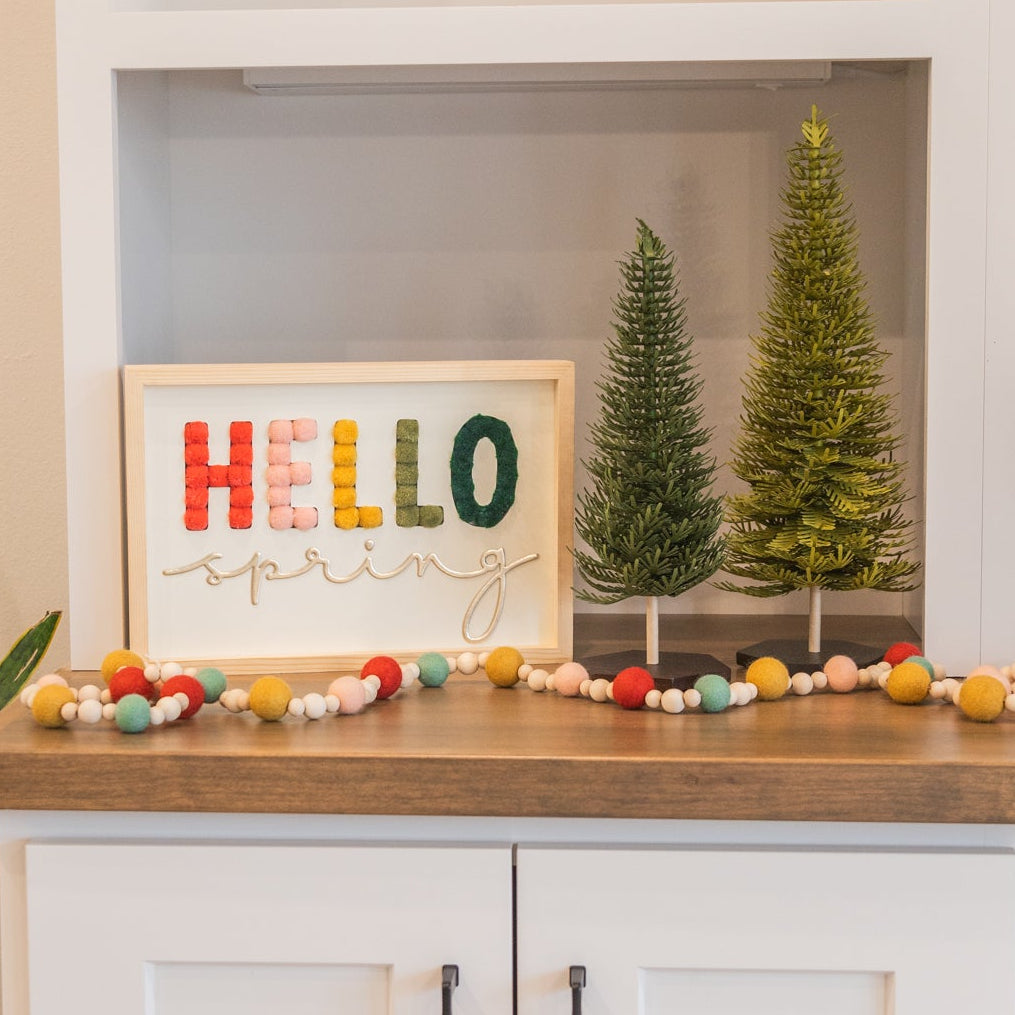 Decorative setup with a 'HELLO SPRING' sign, small Christmas trees, and colorful pom-pom garland on a white cabinet.