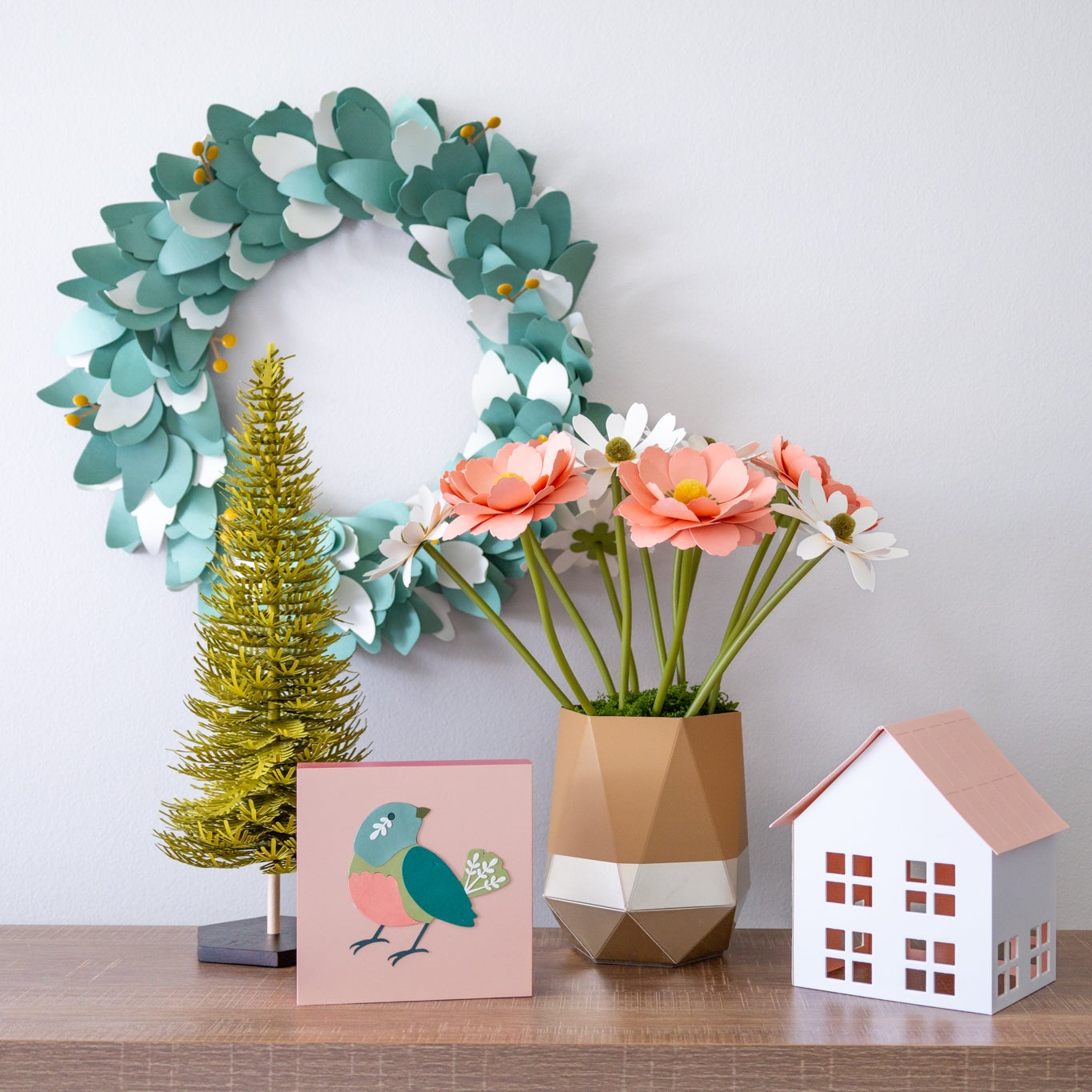 Decorative paper craft items including a wreath, small tree, flowers, and house on a shelf against a plain wall.