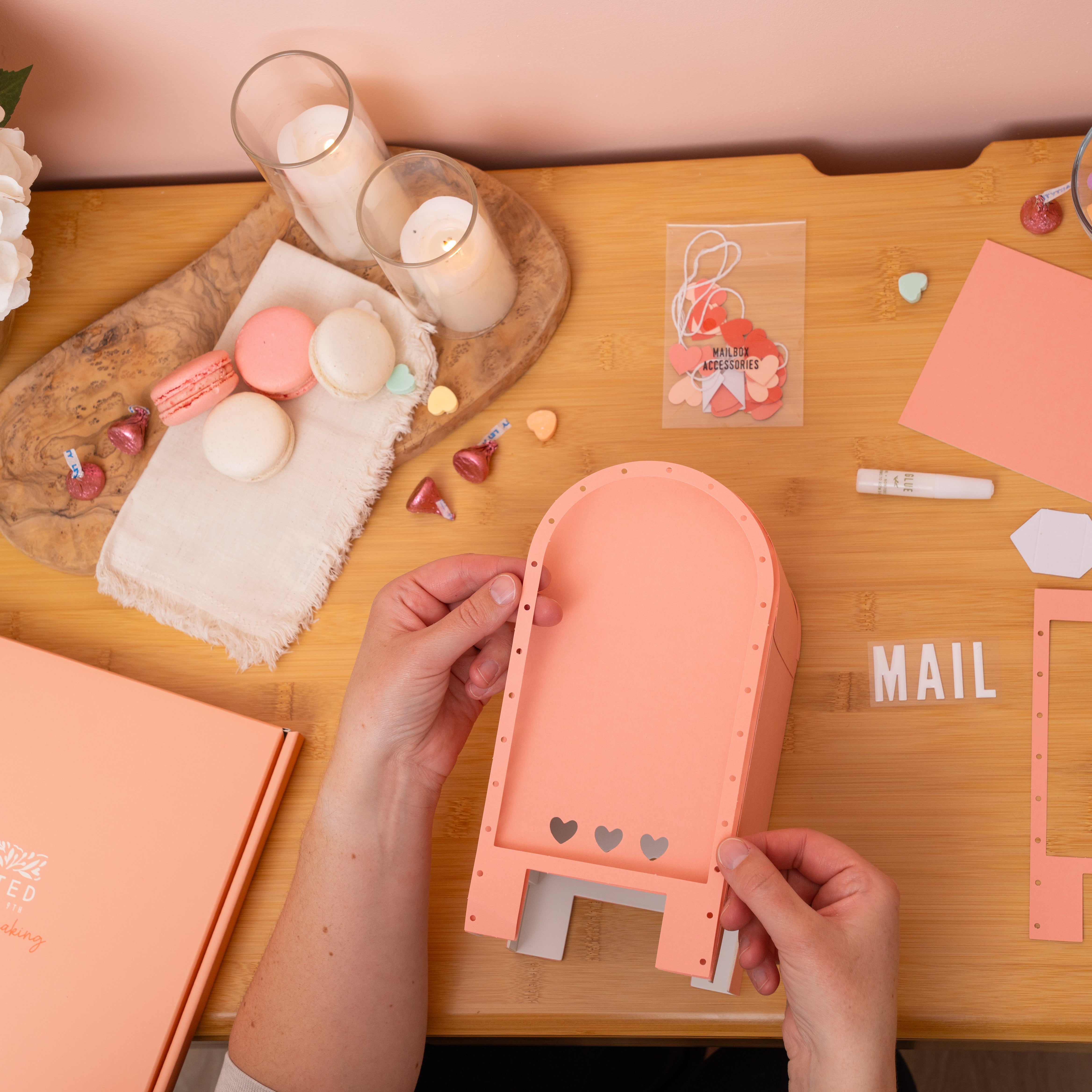 Person assembling a DIY pink letter holder on a wooden table with stationery items.